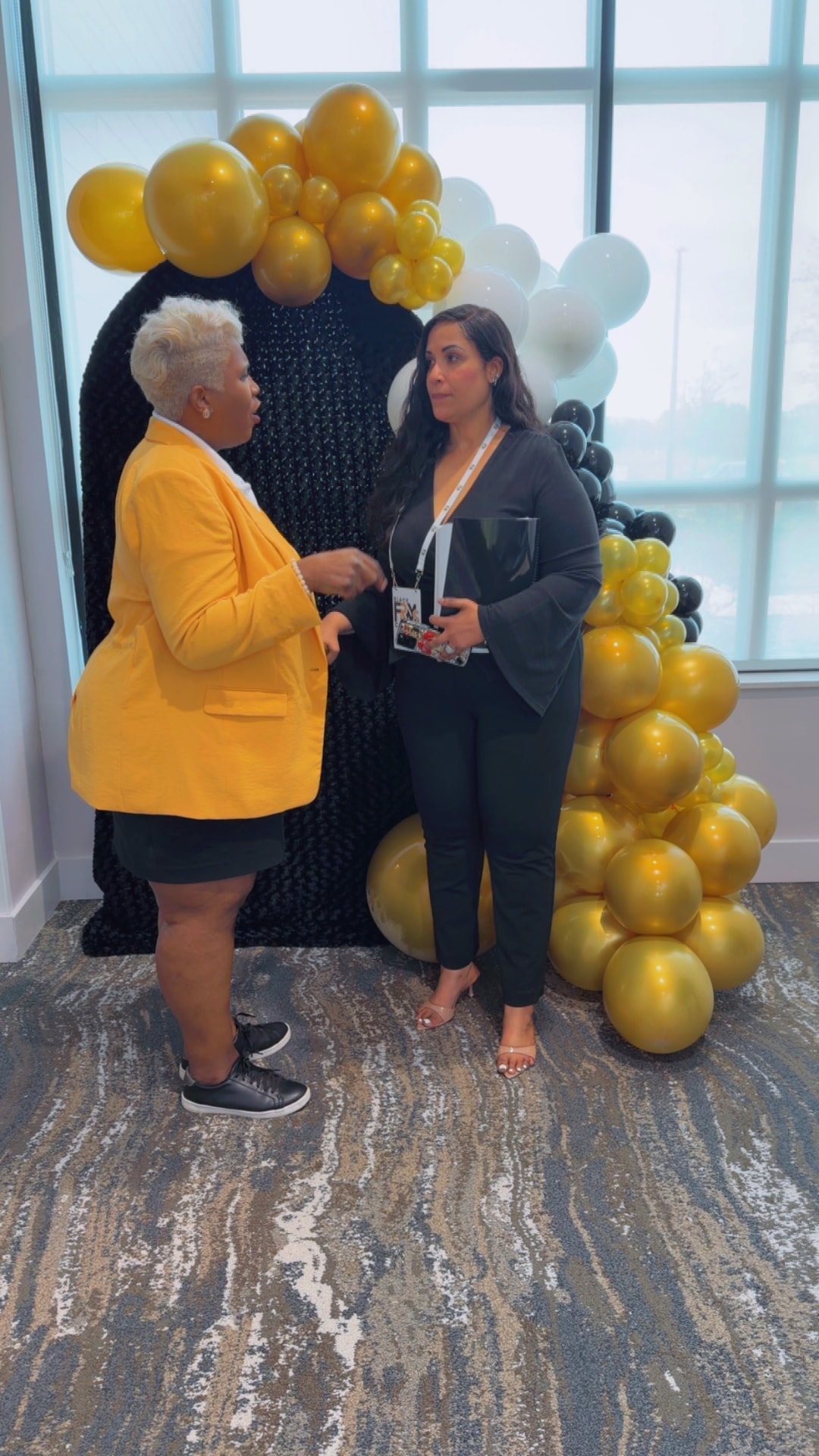 Toya in a gold blazer talking with a member holding a folder at the balloon arch