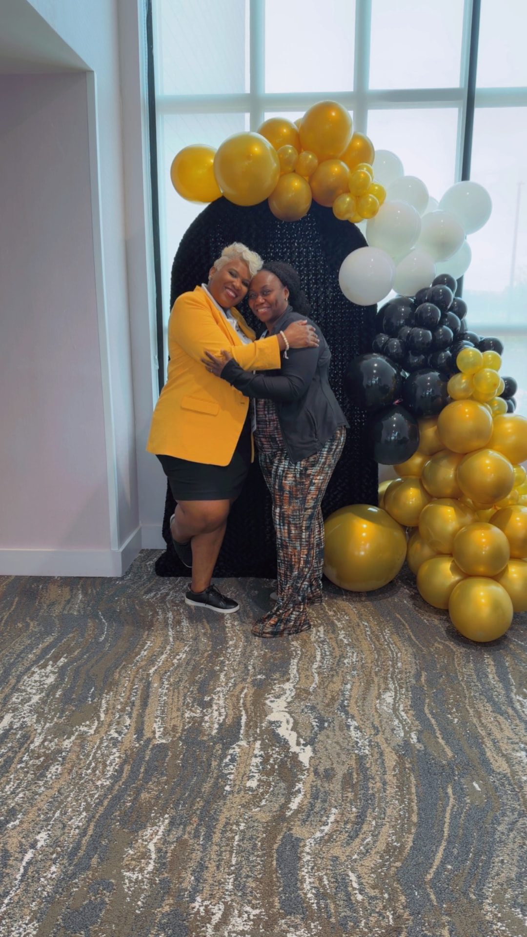 Toya in a gold blazer hugging a member in patterned pants at the balloon arch