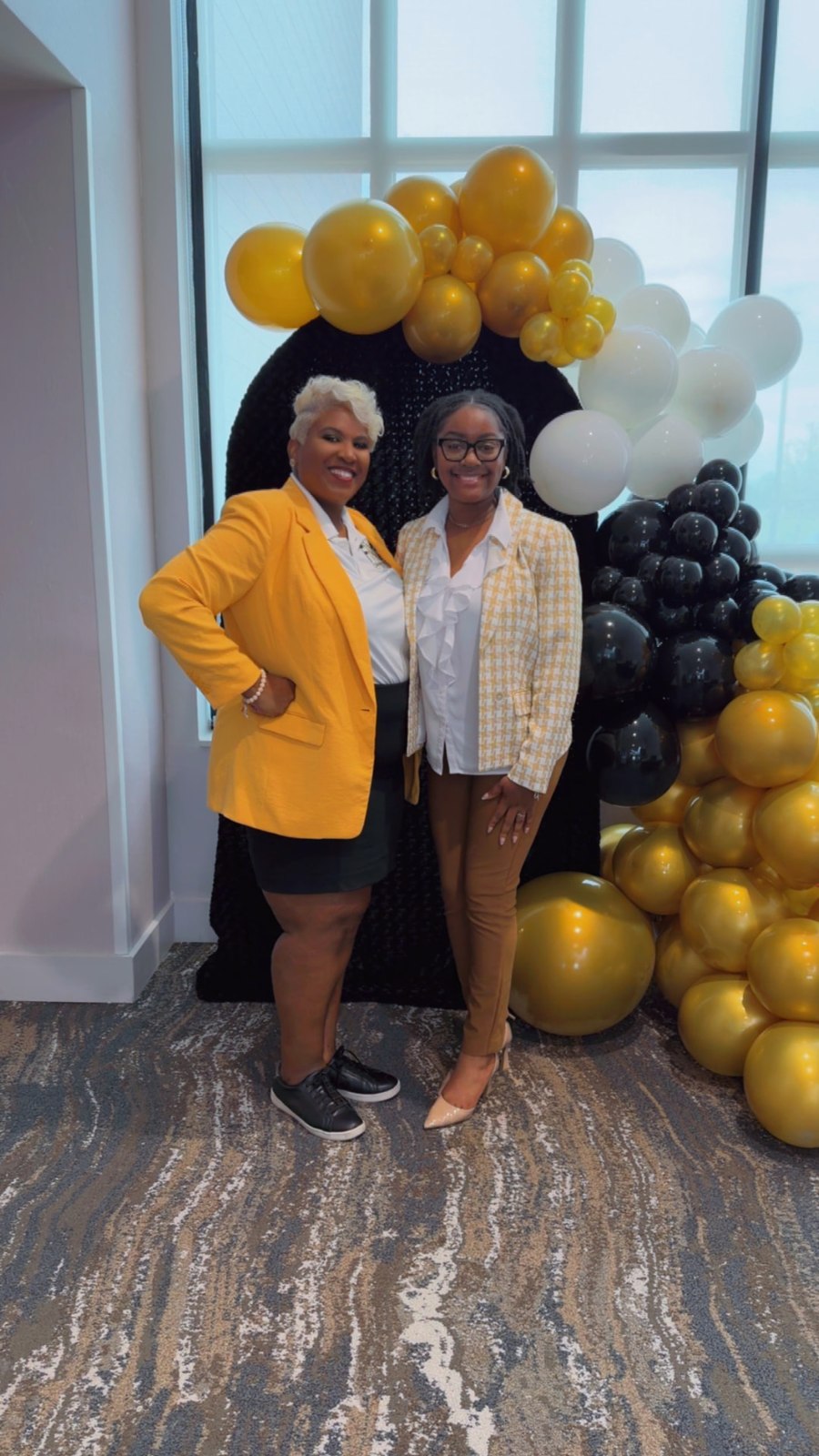 Toya and a member in a patterned blouse smiling beside the balloon arch